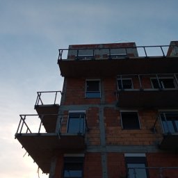 GSM SPOLKA Z OGRANICZONA ODPOWIEDZIALNOSCIA - Surowy budynek wielorodzinny w trakcie budowy, widok z dołu na ceglaną elewację z balkonami z metalowymi balustradami i oknami z roletami, na tle pochmurnego nieba.