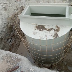 GSM SPOLKA Z OGRANICZONA ODPOWIEDZIALNOSCIA - Cylindryczny, biały zbiornik z pokrywą w wykopie, zabezpieczony siatką z prętów zbrojeniowych, widoczny fragment rury odpływowej.