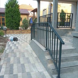 GSM SPOLKA Z OGRANICZONA ODPOWIEDZIALNOSCIA - Metalowa balustrada schodowa w kolorze grafitowym, zamontowana przy wejściu do domu z kostki brukowej, widok z boku, na tle domu z żółtą elewacją i zielenią.