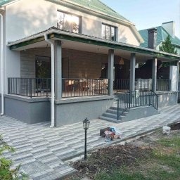 GSM SPOLKA Z OGRANICZONA ODPOWIEDZIALNOSCIA - Nowoczesny dom z szarą elewacją, zielonym dachem i betonowymi schodami z czarną metalową balustradą prowadzącymi na zadaszony taras, widoczne kostki brukowe na podjeździe i lampa ogrodowa.