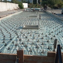 GSM SPOLKA Z OGRANICZONA ODPOWIEDZIALNOSCIA - Przygotowanie zbrojenia płyty fundamentowej: siatka stalowa na podkładkach dystansowych, szalunek drewniany w tle, widoczni pracownicy budowlani.