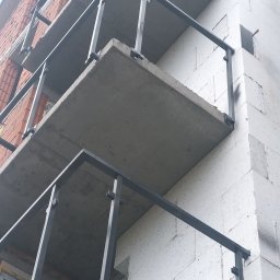 GSM SPOLKA Z OGRANICZONA ODPOWIEDZIALNOSCIA - Surowe betonowe balkony z czarnymi metalowymi balustradami w trakcie budowy, widok z dołu na budynek z białego betonu komórkowego i czerwonej cegły.