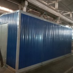 GSM SPOLKA Z OGRANICZONA ODPOWIEDZIALNOSCIA - Kontener biurowy z blachy trapezowej w kolorze niebieskim z szarymi ramami i drzwiami, umieszczony w hali produkcyjnej, z widoczną osobą w lewym dolnym rogu.