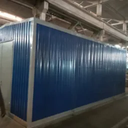 Kontener biurowy z blachy trapezowej w kolorze niebieskim z szarymi ramami i drzwiami, umieszczony w hali produkcyjnej, z widoczną osobą w lewym dolnym rogu.