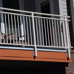 GSM SPOLKA Z OGRANICZONA ODPOWIEDZIALNOSCIA - Biała metalowa balustrada balkonu z widocznymi krzesłami w tle, zamontowana na brązowej konstrukcji, z szarym budynkiem w tle.