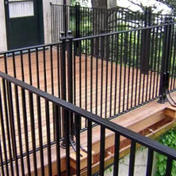 Czarna, metalowa balustrada z dekoracyjnymi kulami na słupkach, otaczająca drewniany taras na zewnątrz budynku z zielonymi roślinami w tle.