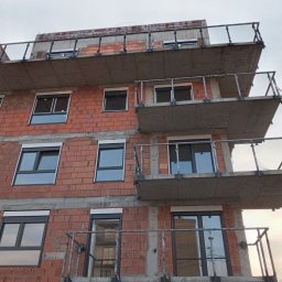 GSM SPOLKA Z OGRANICZONA ODPOWIEDZIALNOSCIA - Budynek wielorodzinny w trakcie budowy, widoczne surowe ściany z czerwonej cegły, zamontowane okna z roletami zewnętrznymi i balkony z metalowymi balustradami, betonowe płyty balkonowe.