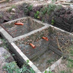 GSM SPOLKA Z OGRANICZONA ODPOWIEDZIALNOSCIA - Dwa połączone murem zbiorniki z cegły z wystającymi rurami kanalizacyjnymi, wykopane w ziemi, widoczne ślady zaprawy.