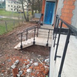 GSM SPOLKA Z OGRANICZONA ODPOWIEDZIALNOSCIA - Zewnętrzne schody betonowe z czarną metalową balustradą prowadzące do wejścia zabezpieczonego niebieską folią ochronną, teren wokół w trakcie prac wykończeniowych z gruzem i resztkami cegieł.