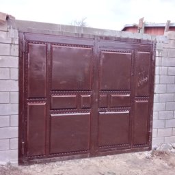 GSM SPOLKA Z OGRANICZONA ODPOWIEDZIALNOSCIA - Brązowa, metalowa brama dwuskrzydłowa z dekoracyjnymi panelami, osadzona w murze z szarych bloczków betonowych, z widocznymi śladami zaprawy i drewnianymi elementami na górze.