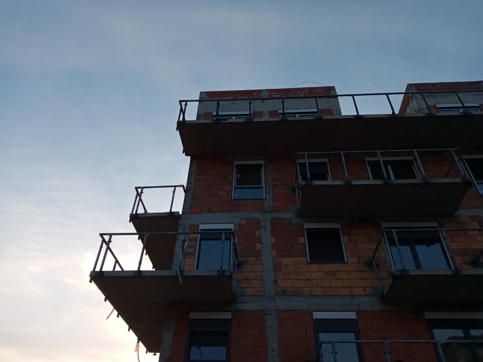 Surowy budynek wielorodzinny w trakcie budowy, widok z dołu na ceglaną elewację z balkonami z metalowymi balustradami i oknami z roletami, na tle pochmurnego nieba.