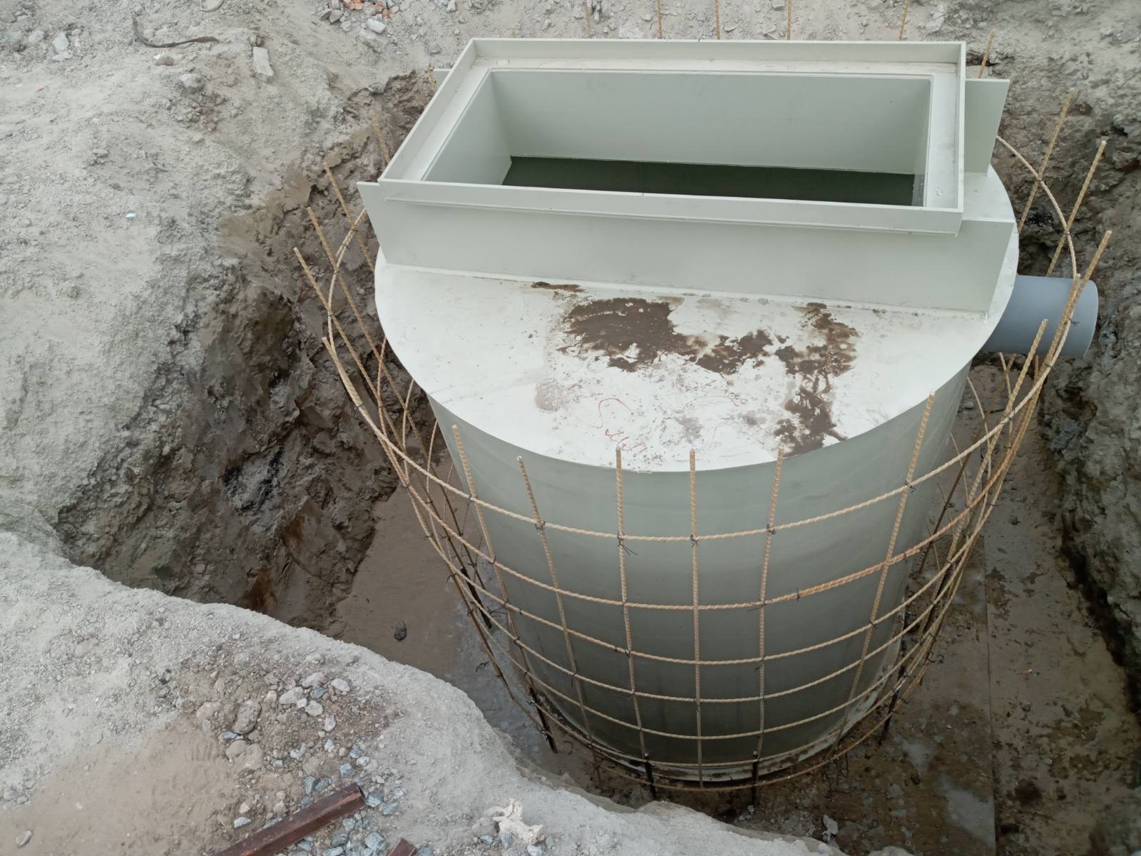 Cylindryczny, biały zbiornik z pokrywą w wykopie, zabezpieczony siatką z prętów zbrojeniowych, widoczny fragment rury odpływowej.