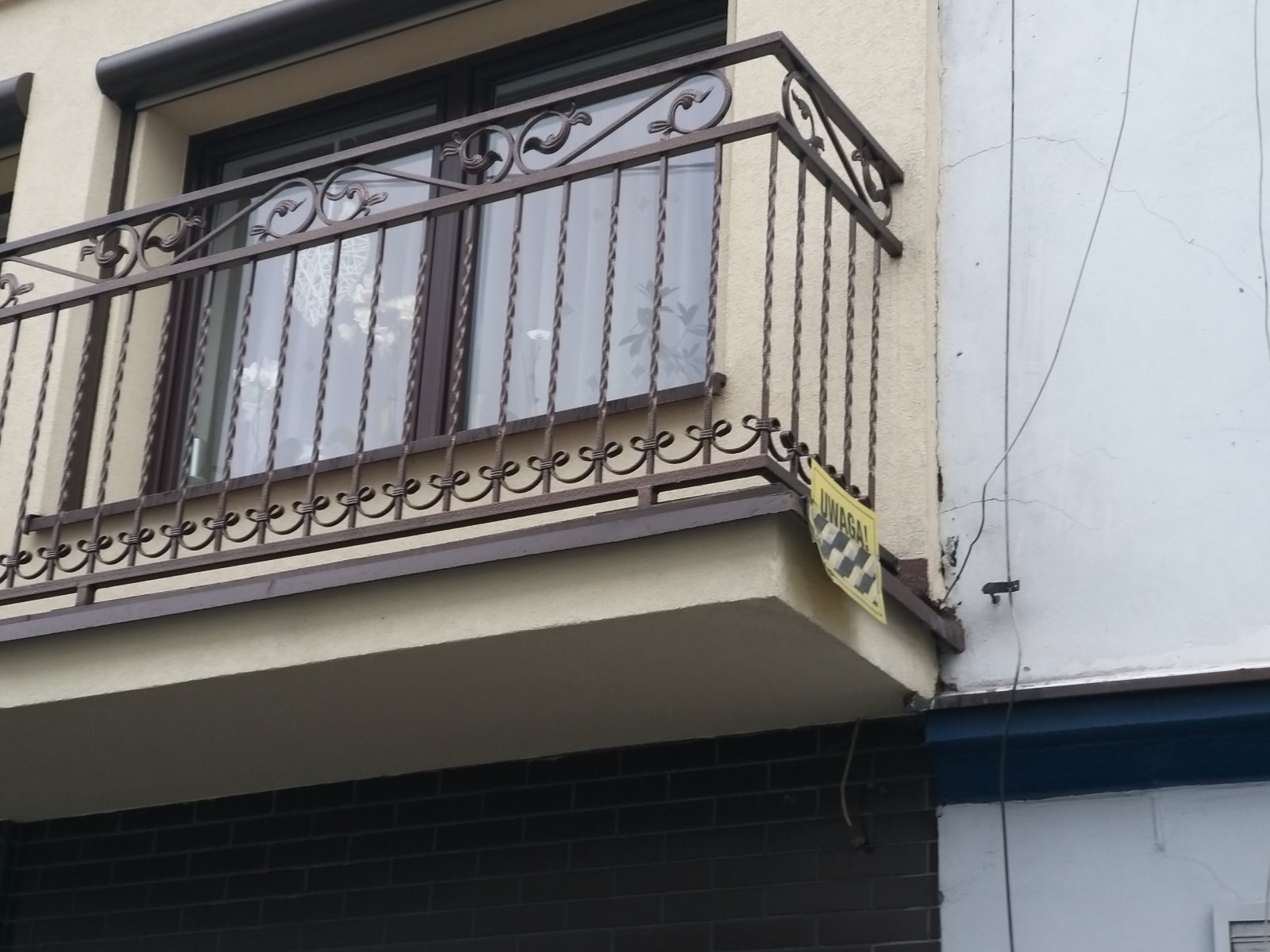 Kuty balkon z ozdobnymi elementami i żółtą tabliczką ostrzegawczą przymocowaną do barierki, widok z dołu.