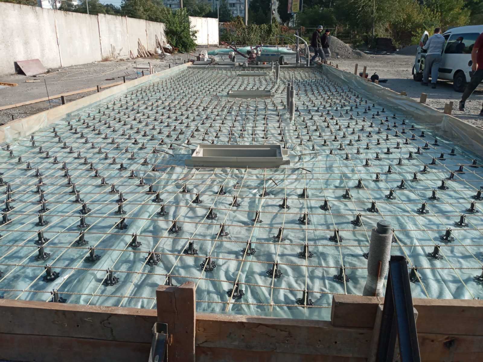 Przygotowanie zbrojenia płyty fundamentowej: siatka stalowa na podkładkach dystansowych, szalunek drewniany w tle, widoczni pracownicy budowlani.