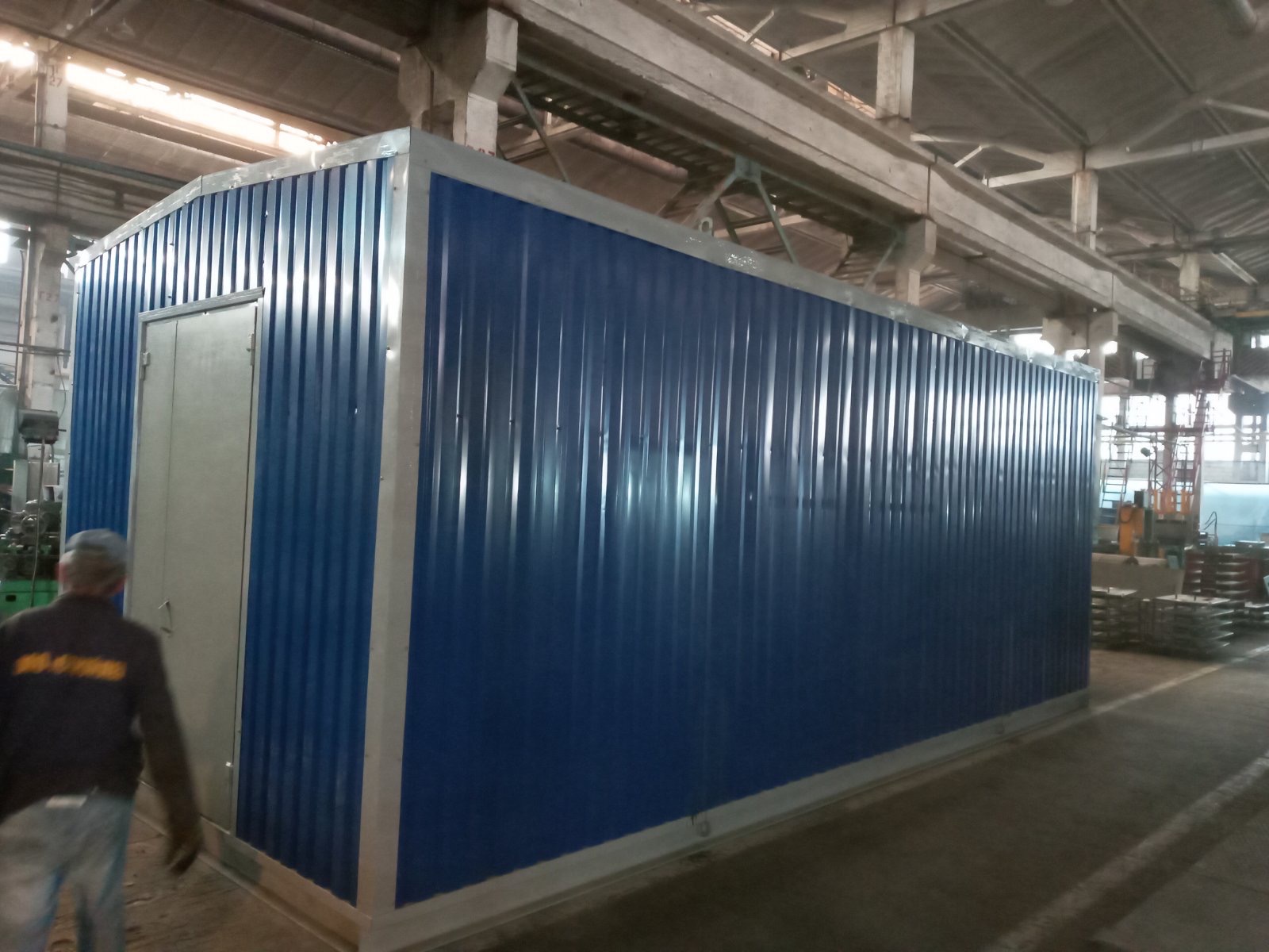 Kontener biurowy z blachy trapezowej w kolorze niebieskim z szarymi ramami i drzwiami, umieszczony w hali produkcyjnej, z widoczną osobą w lewym dolnym rogu.