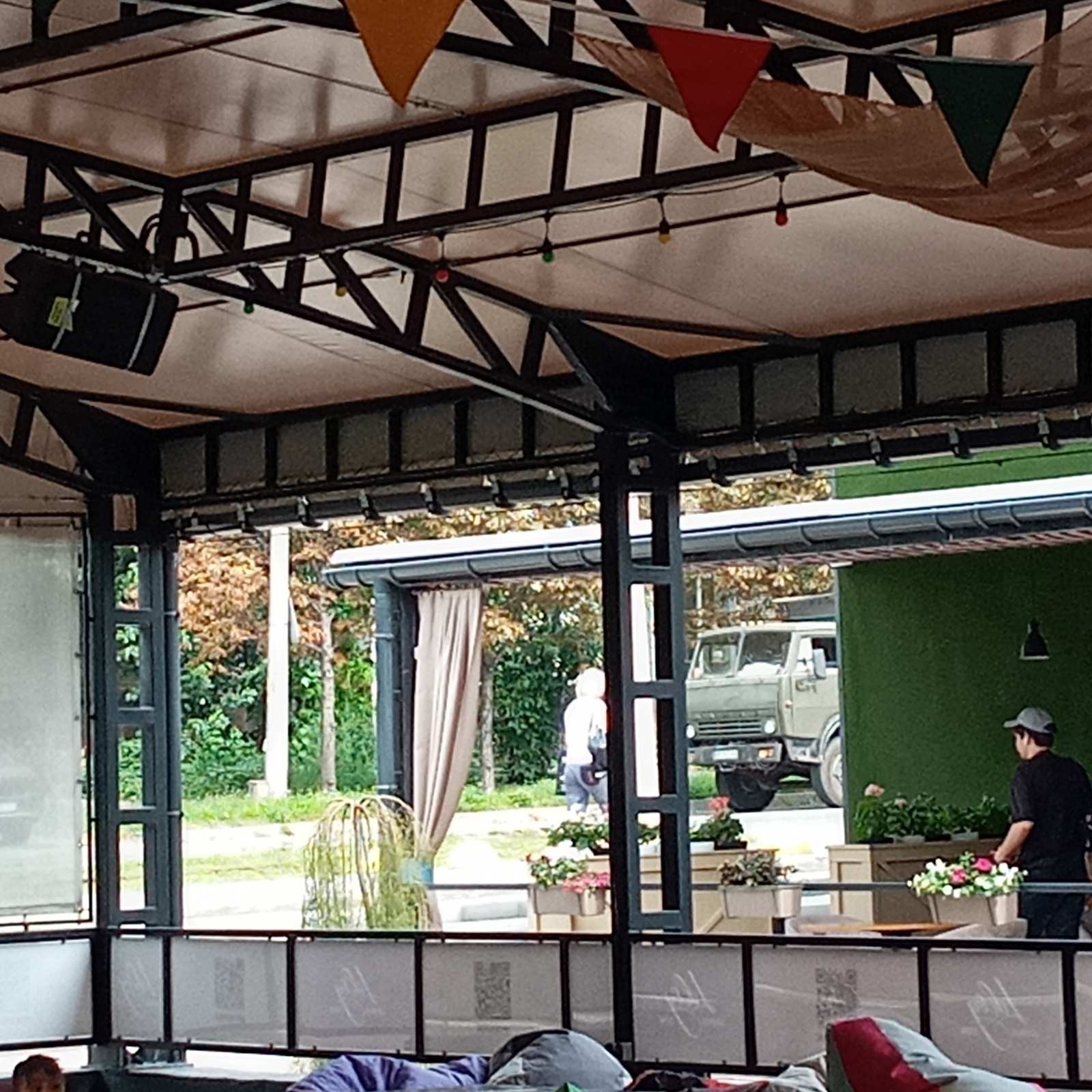 Metalowa konstrukcja pergoli z dekoracyjnymi trójkątnymi flagami i oświetleniem, widok na zewnątrz z roślinnością, samochodem i osobami.