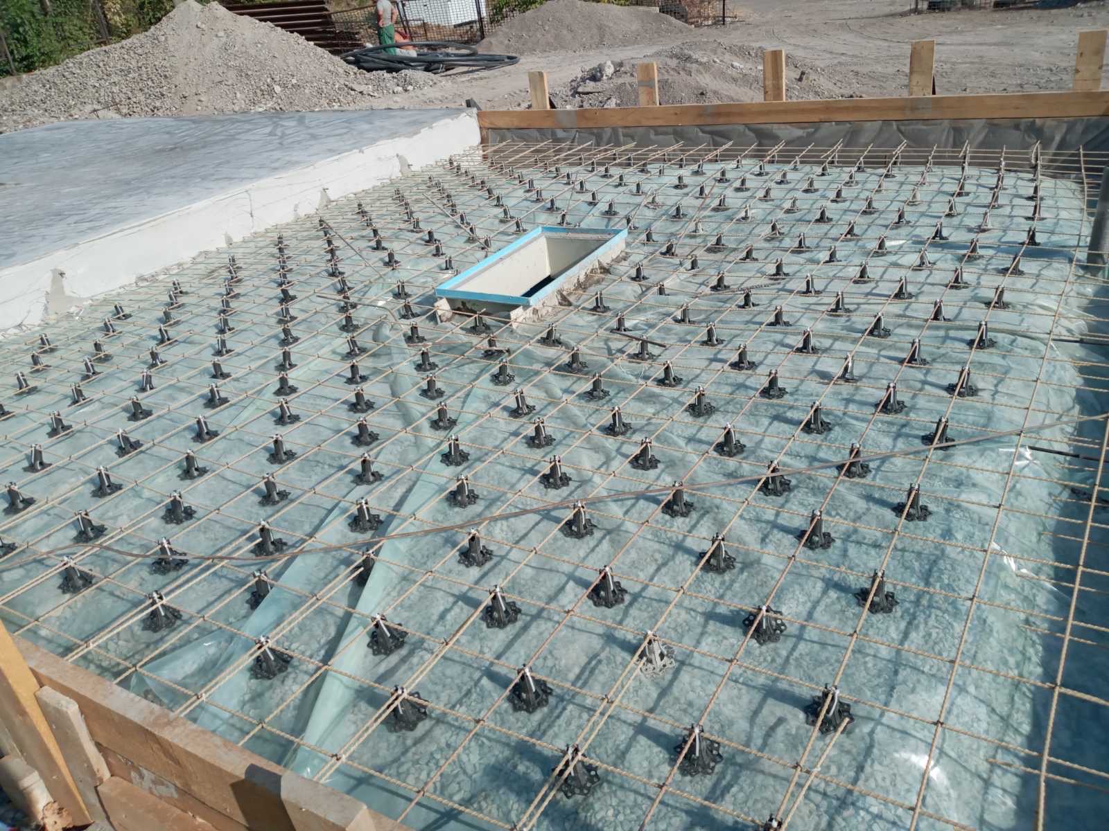 Przygotowanie zbrojenia płyty fundamentowej z użyciem dystansów pod siatką, widoczny szalunek i wykop pod studzienkę rewizyjną.