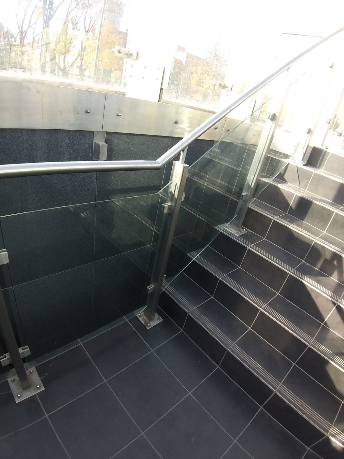 Schody wyłożone czarnymi płytkami, z metalową poręczą i szklanym panelem ochronnym, widoczne elementy mocujące.