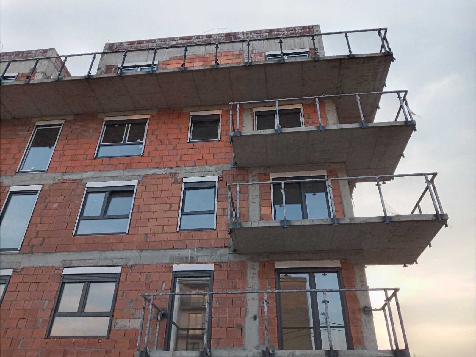 Budynek wielorodzinny w trakcie budowy, widoczne surowe ściany z czerwonej cegły, zamontowane okna z roletami zewnętrznymi i balkony z metalowymi balustradami, betonowe płyty balkonowe.