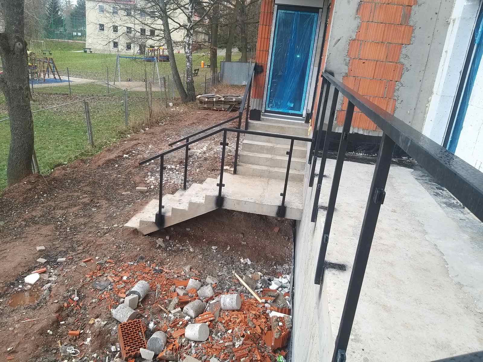 Zewnętrzne schody betonowe z czarną metalową balustradą prowadzące do wejścia zabezpieczonego niebieską folią ochronną, teren wokół w trakcie prac wykończeniowych z gruzem i resztkami cegieł.