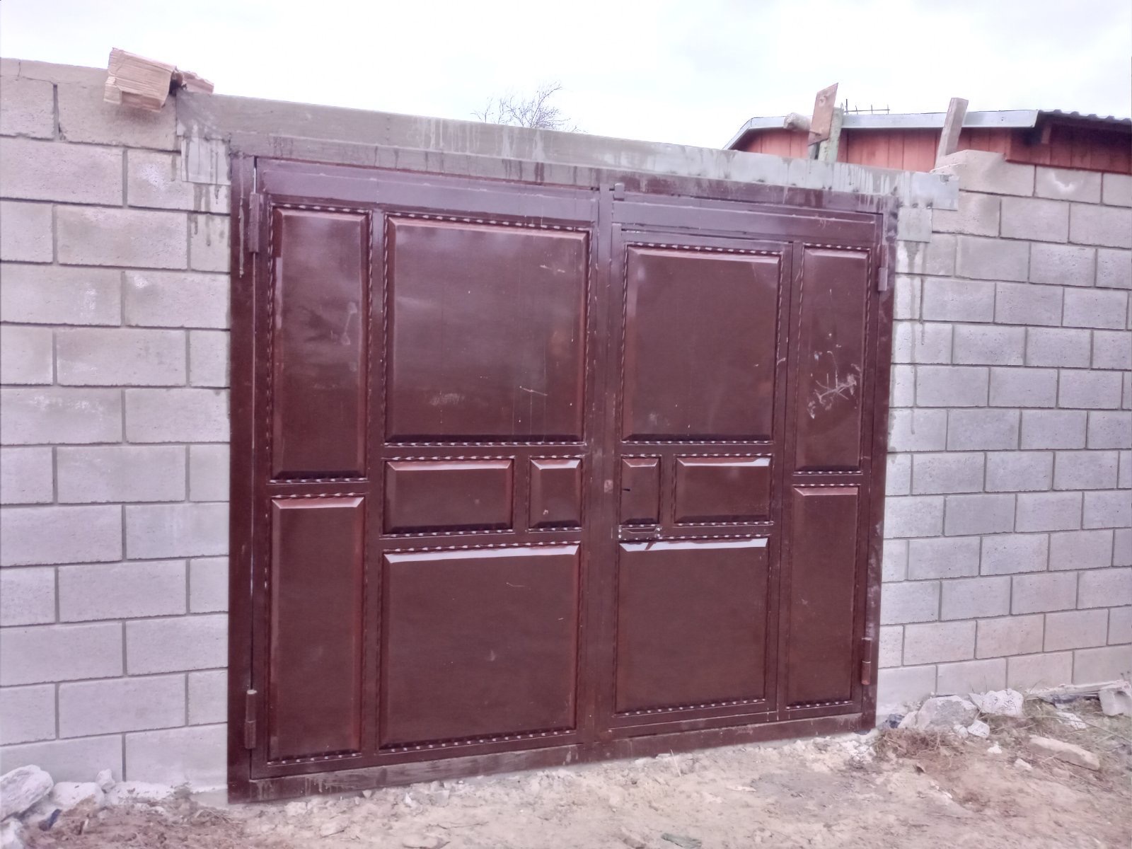 Brązowa, metalowa brama dwuskrzydłowa z dekoracyjnymi panelami, osadzona w murze z szarych bloczków betonowych, z widocznymi śladami zaprawy i drewnianymi elementami na górze.