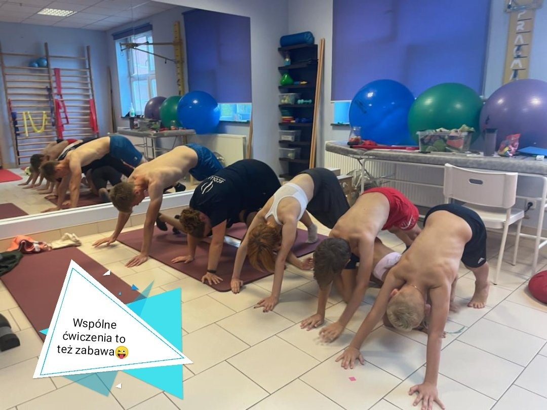 Grupa osób w różnym wieku ćwiczy w sali gimnastycznej, wykonując pozycję psa z głową w dół na matach do ćwiczeń. W tle widoczne lustro, drabinki gimnastyczne, kolorowe piłki i półki z akcesoriami.