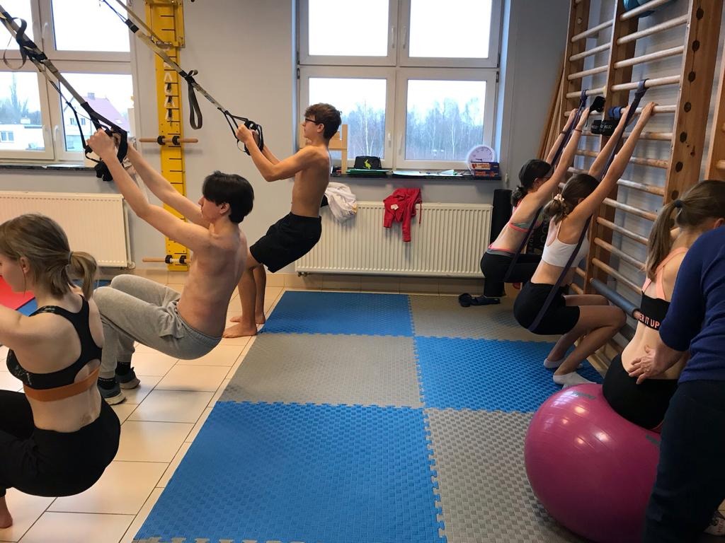 Grupa osób ćwiczy w sali rehabilitacyjnej z wykorzystaniem sprzętu do ćwiczeń, w tym TRX i drabinek gimnastycznych, na podłodze z puzzli piankowych.