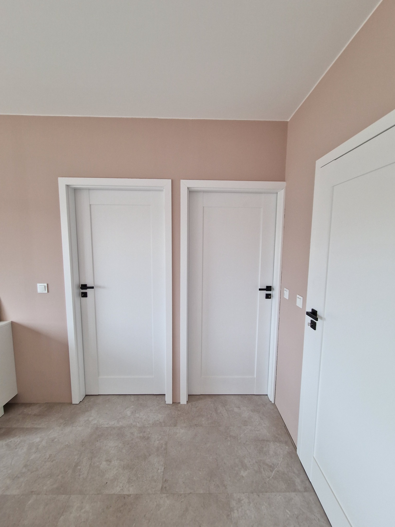 Wnętrze z trzema białymi, minimalistycznymi drzwiami z czarnymi klamkami na tle ścian w odcieniach beżu i szarości, widoczne płytki podłogowe imitujące kamień.