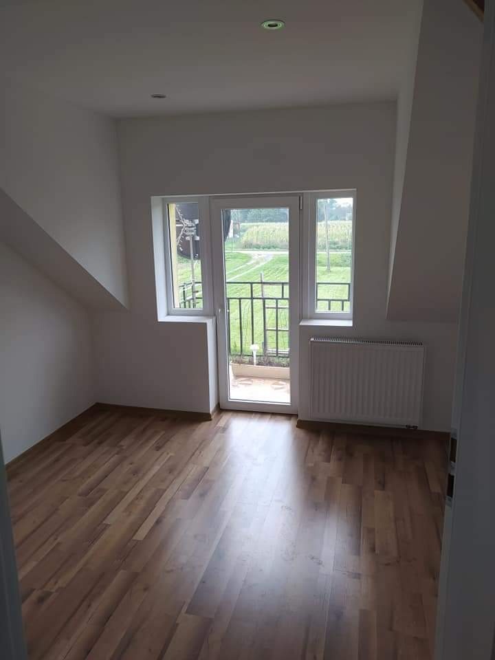 Poddasze w stanie deweloperskim z drewnopodobną podłogą, białymi ścianami, grzejnikiem i oknem balkonowym z widokiem na zieleń.