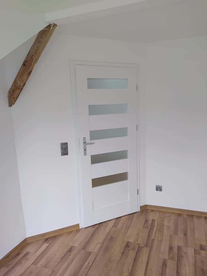 Wnętrze poddasza z białą ścianą, drewnianą belką stropową i drzwiami z przeszkleniami, widoczna podłoga z paneli imitujących drewno.