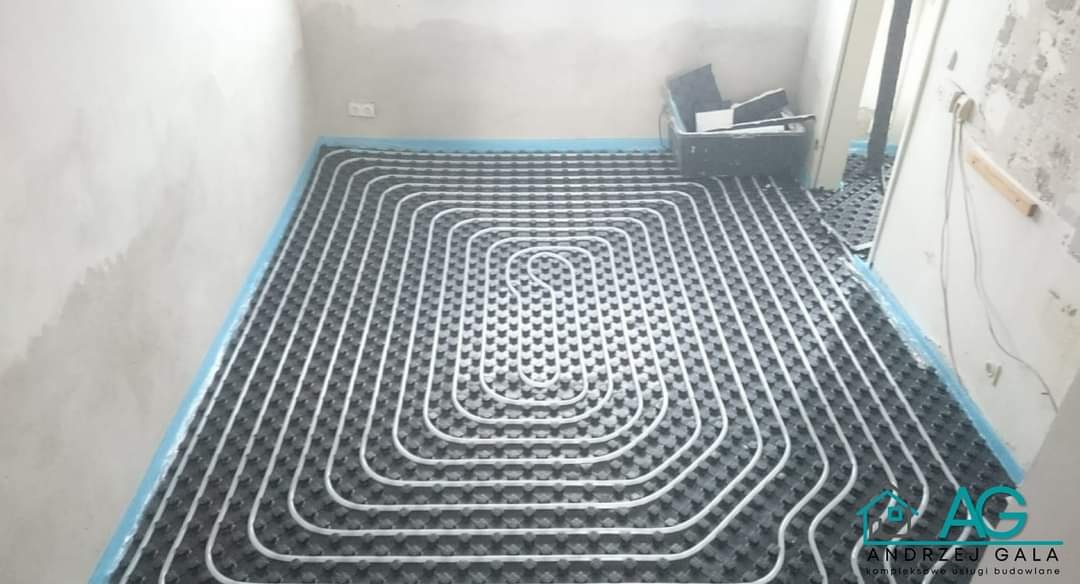 Rozłożone rury ogrzewania podłogowego na czarnej macie izolacyjnej w surowym pomieszczeniu, zabezpieczone niebieską folią przy ścianach.