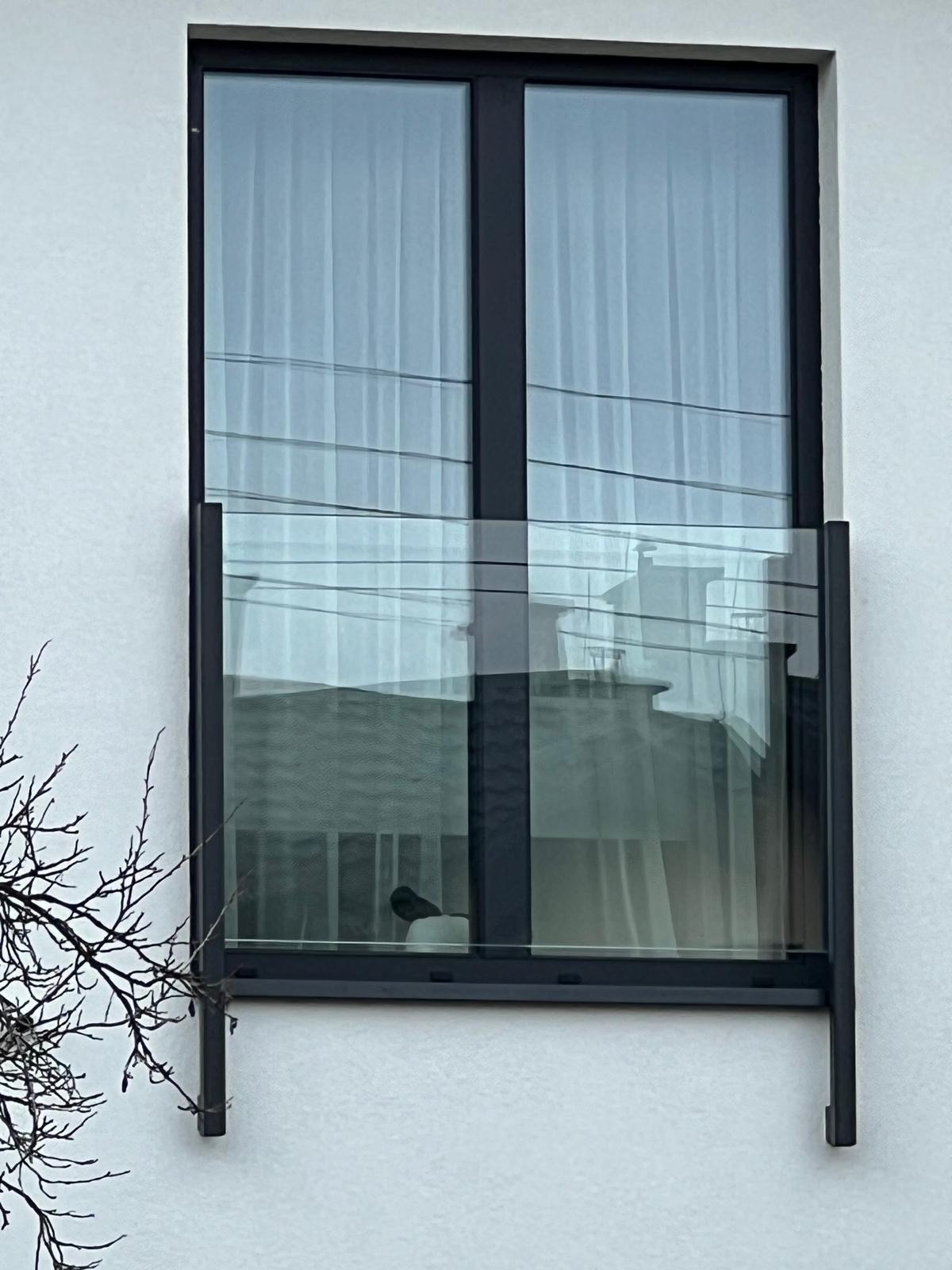 Szklana balustrada balkonowa z czarną ramą, widoczna z zewnątrz budynku. Minimalistyczny design, odbicie otoczenia w szkle. Gałąź drzewa w lewym dolnym rogu.