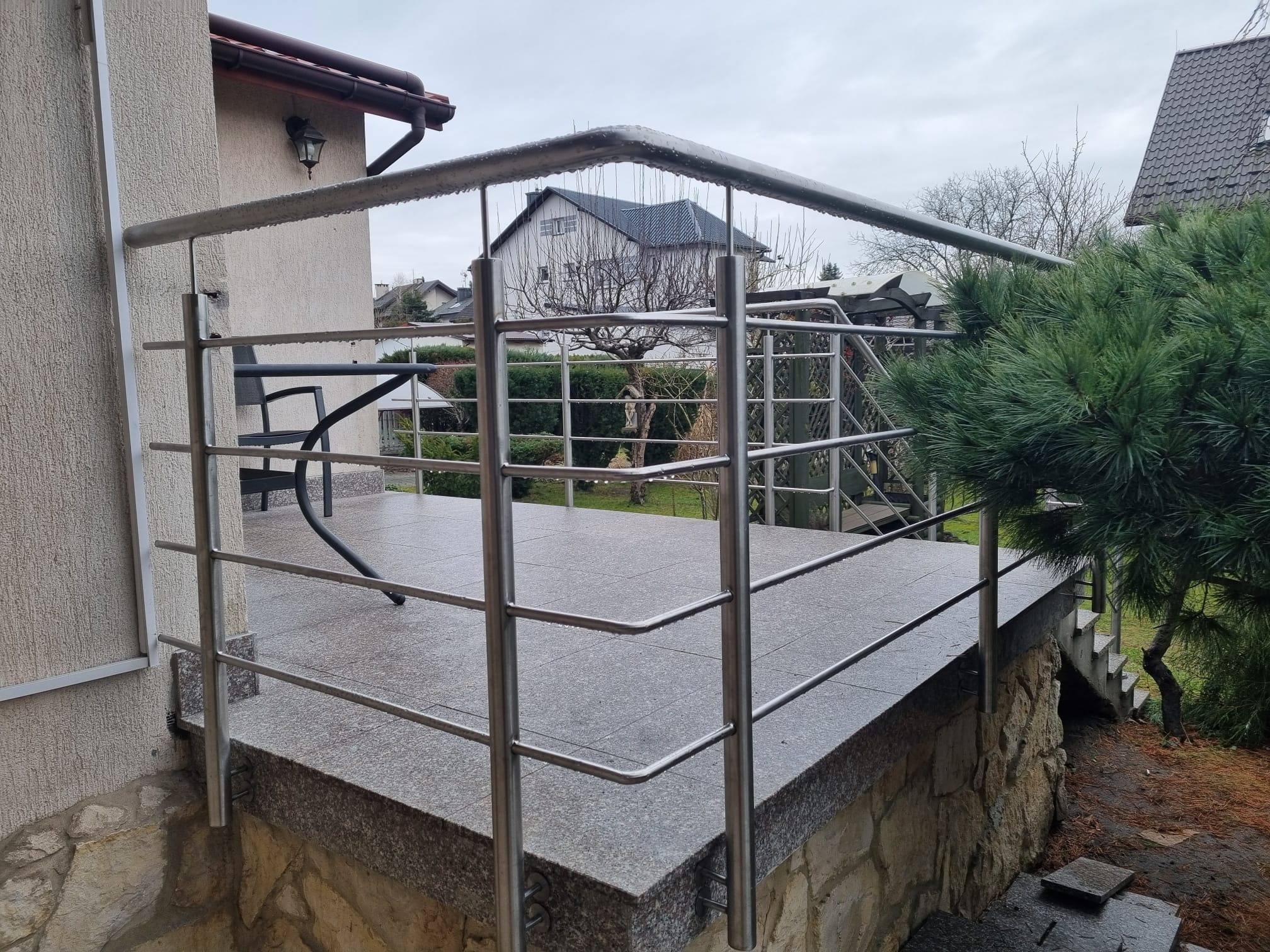 Nowoczesna balustrada nierdzewna z poziomymi prętami na tarasie wykończonym kamieniem, widok na ogród i dom w tle, pochmurny dzień.
