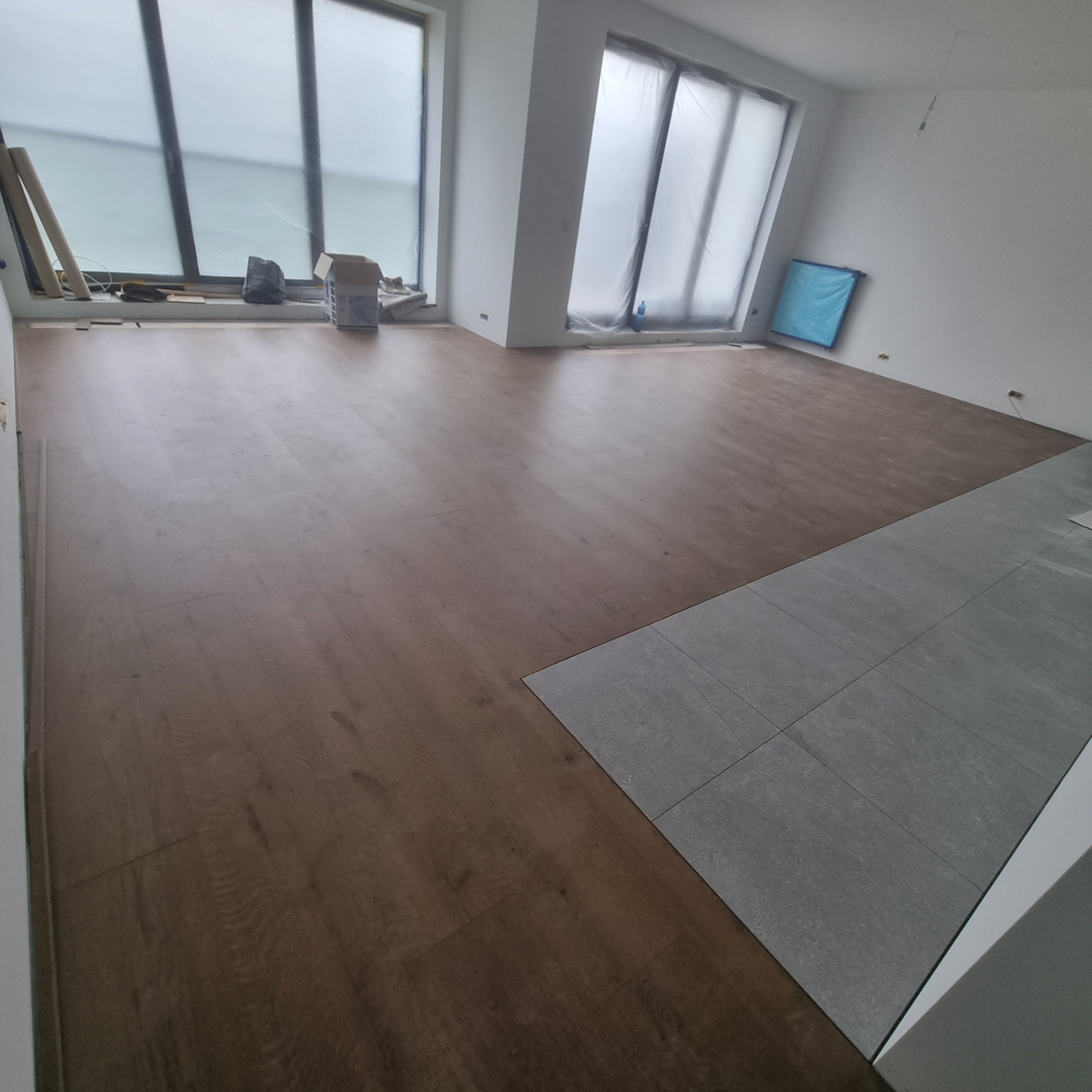 Układanie paneli podłogowych w nowym mieszkaniu, widoczne panele w kolorze ciemnego drewna i fragment ułożonych szarych płytek, okna zabezpieczone folią malarską.