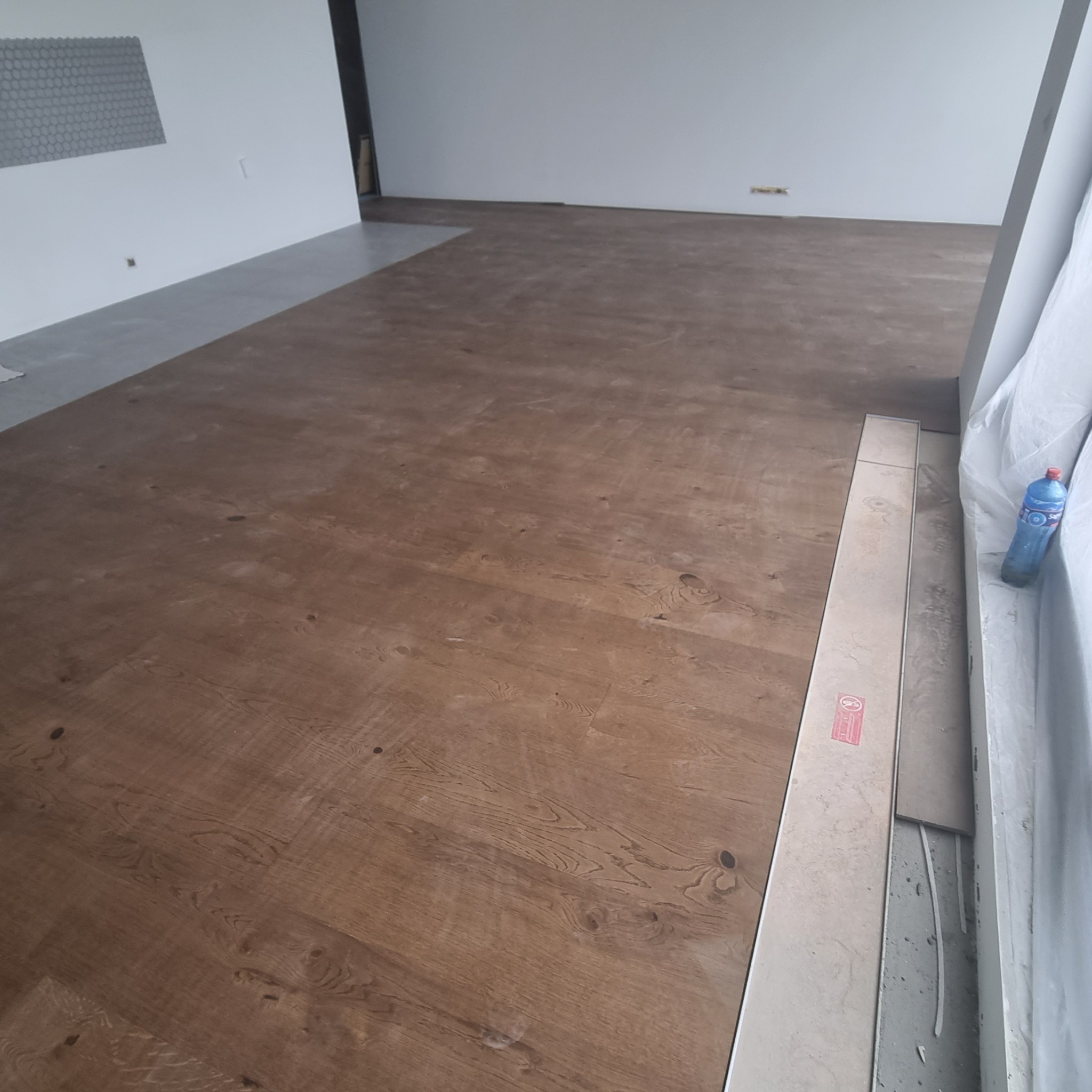 Ułożona podłoga z paneli imitujących drewno w jasnym wnętrzu, widoczne niedociągnięcia przy ścianie i resztki materiałów budowlanych.
