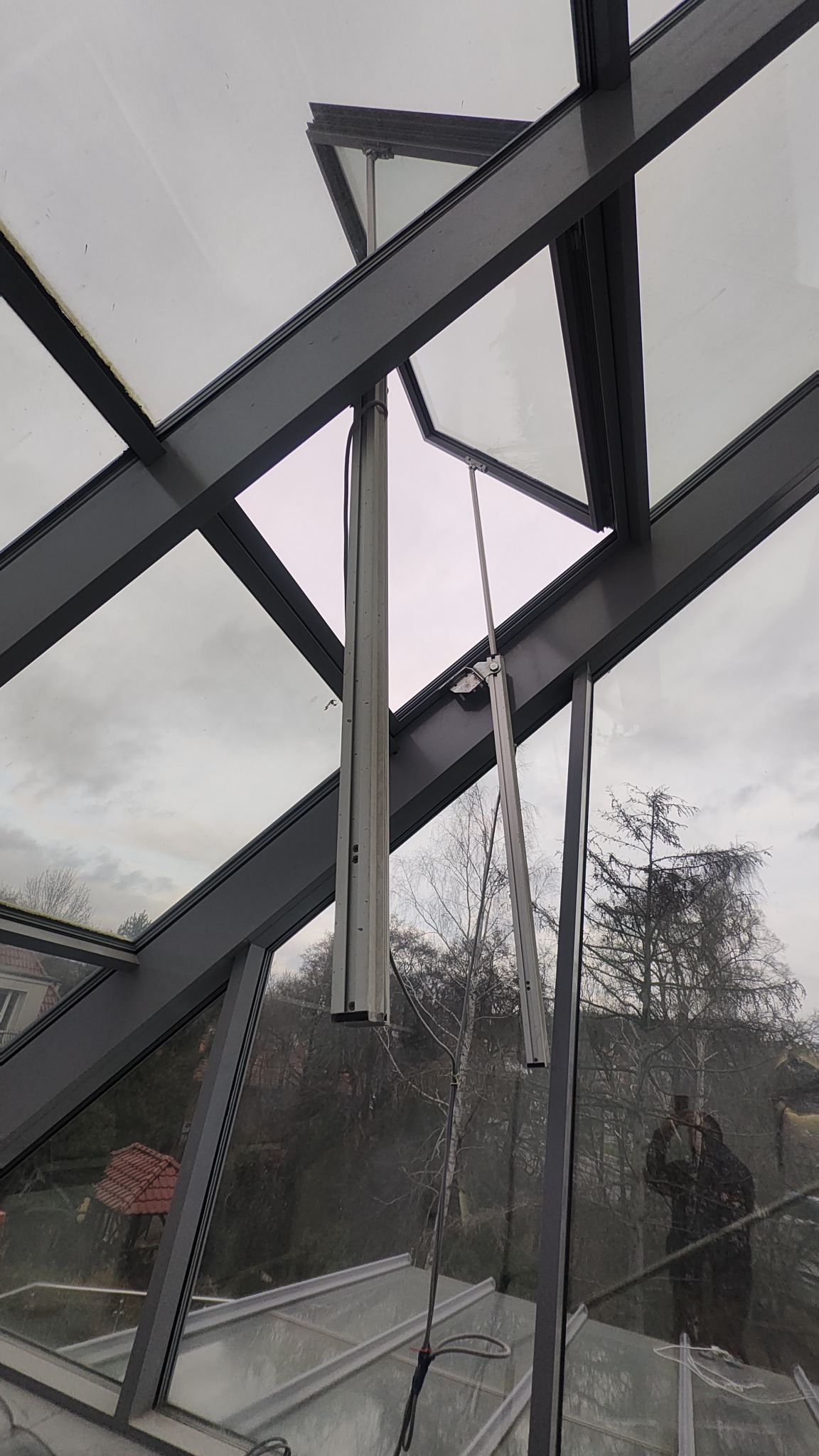 Szklany dach z otwartymi oknami uchylnymi, widoczne mechanizmy otwierania, na zewnątrz drzewa bez liści i budynki.