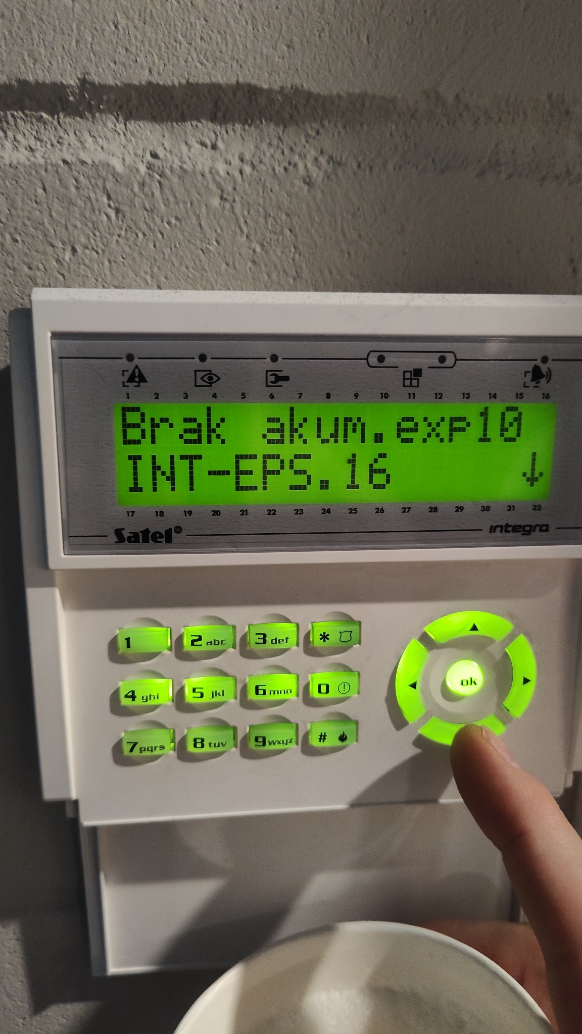 Panel sterowania alarmu Satel Integra z komunikatem o braku akumulatora, podświetlone przyciski i palec naciskający przycisk OK.