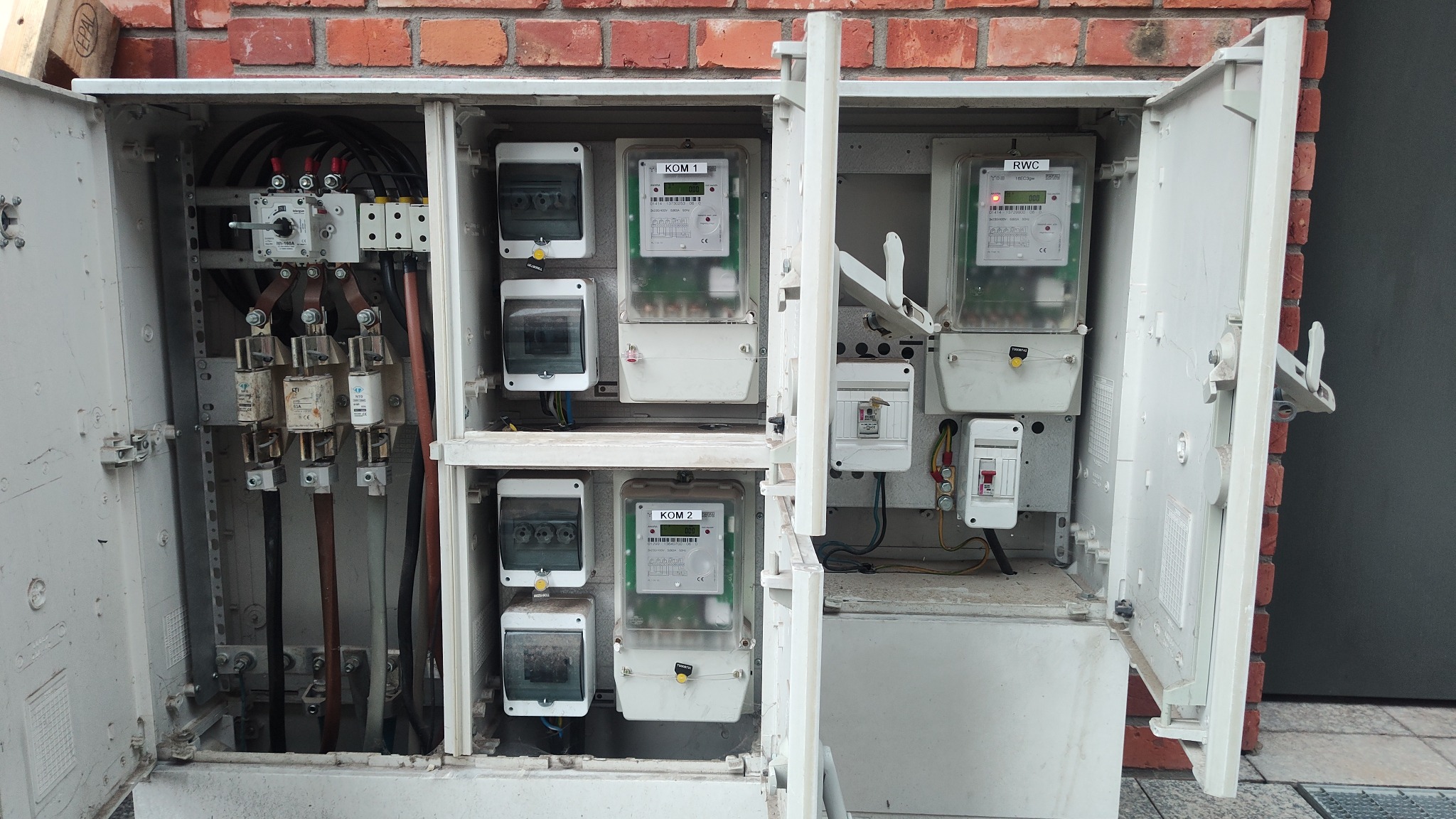 Otwarta skrzynka elektryczna z kilkoma licznikami energii i bezpiecznikami, widoczne przewody i elementy instalacji, tło z ceglanej ściany.