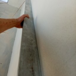 Jarosław Majchrowicz - Ręka przytrzymuje metalowy profil przy ścianie przygotowanej do prac wykończeniowych, widoczny fragment pomieszczenia w trakcie remontu.