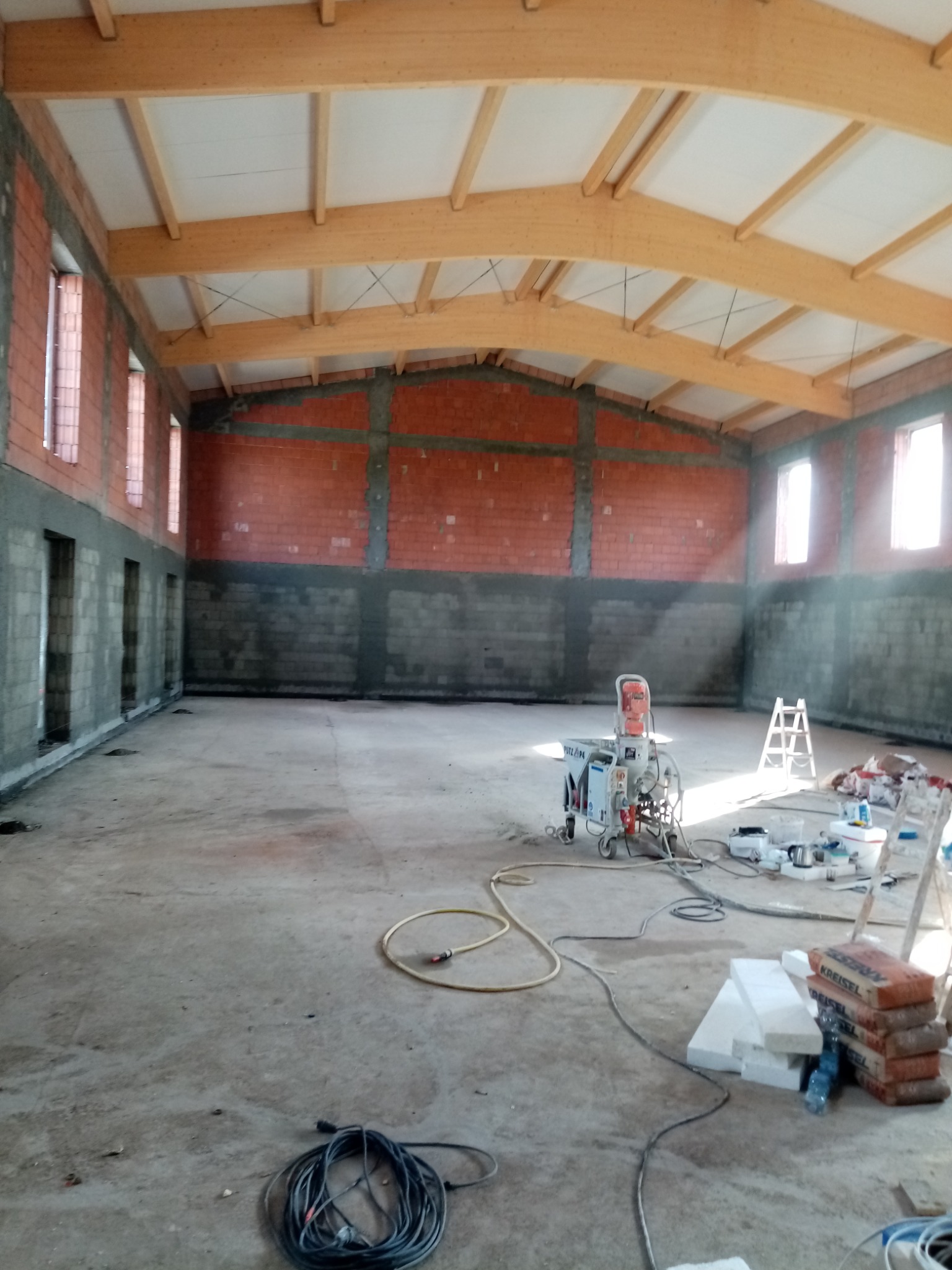 Wnętrze hali w Śremie w trakcie tynkowania maszynowego, widoczna maszyna do tynkowania, ceglane ściany częściowo pokryte tynkiem, drewniana konstrukcja dachu i porozrzucane materiały budowlane.