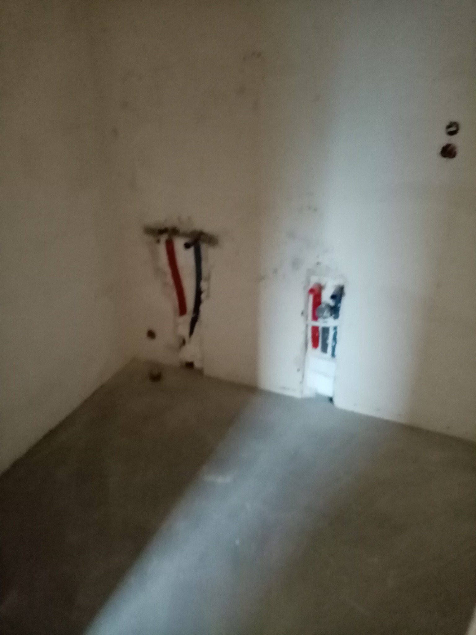 Surowe wnętrze pokoju przed wykończeniem: widoczne instalacje wodne i elektryczne w ścianach, świeżo wylana, szara posadzka.