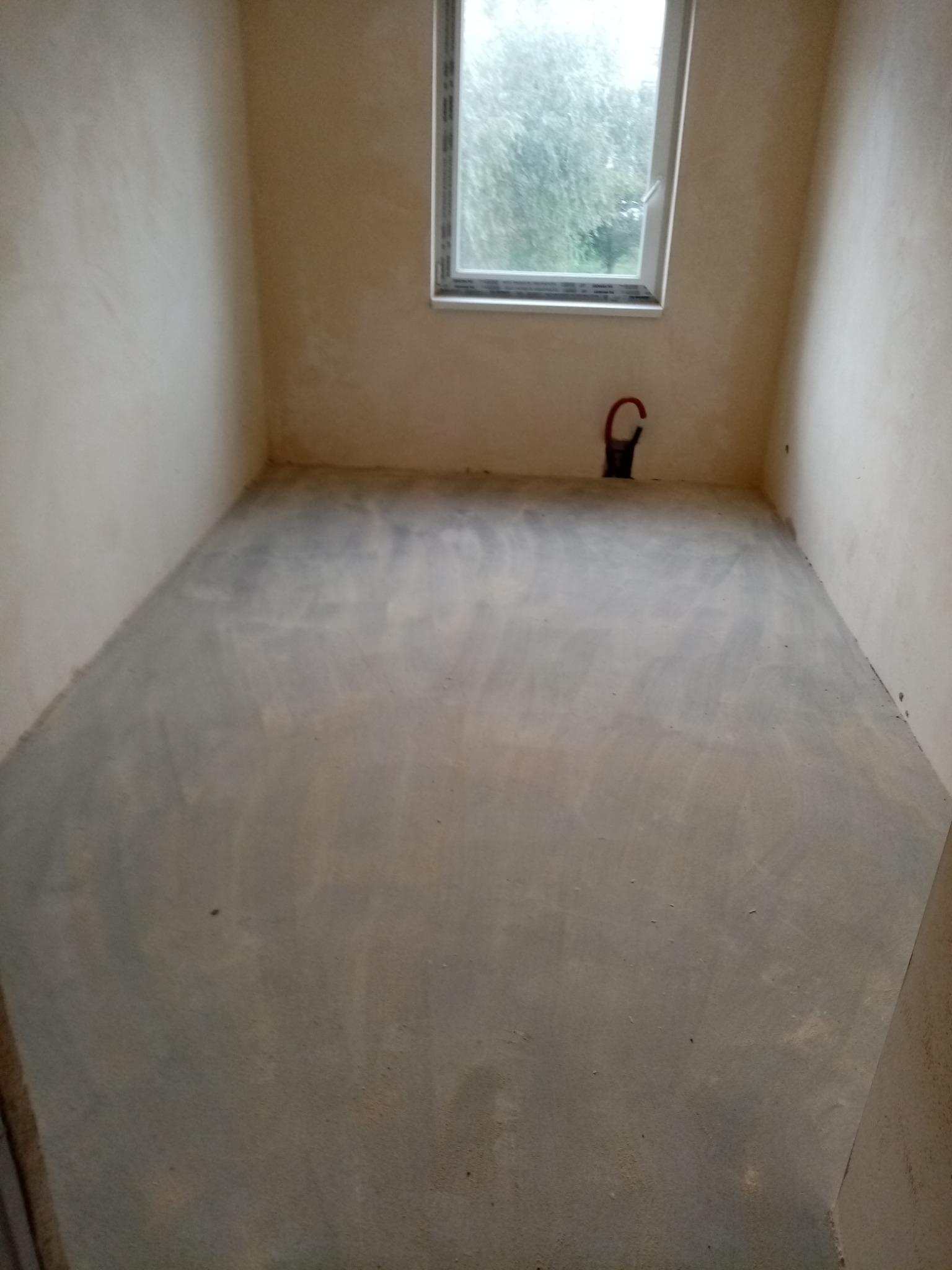 Pokój po wylewce podłogi, ściany przygotowane pod malowanie, widoczne rury instalacji grzewczej przy oknie.