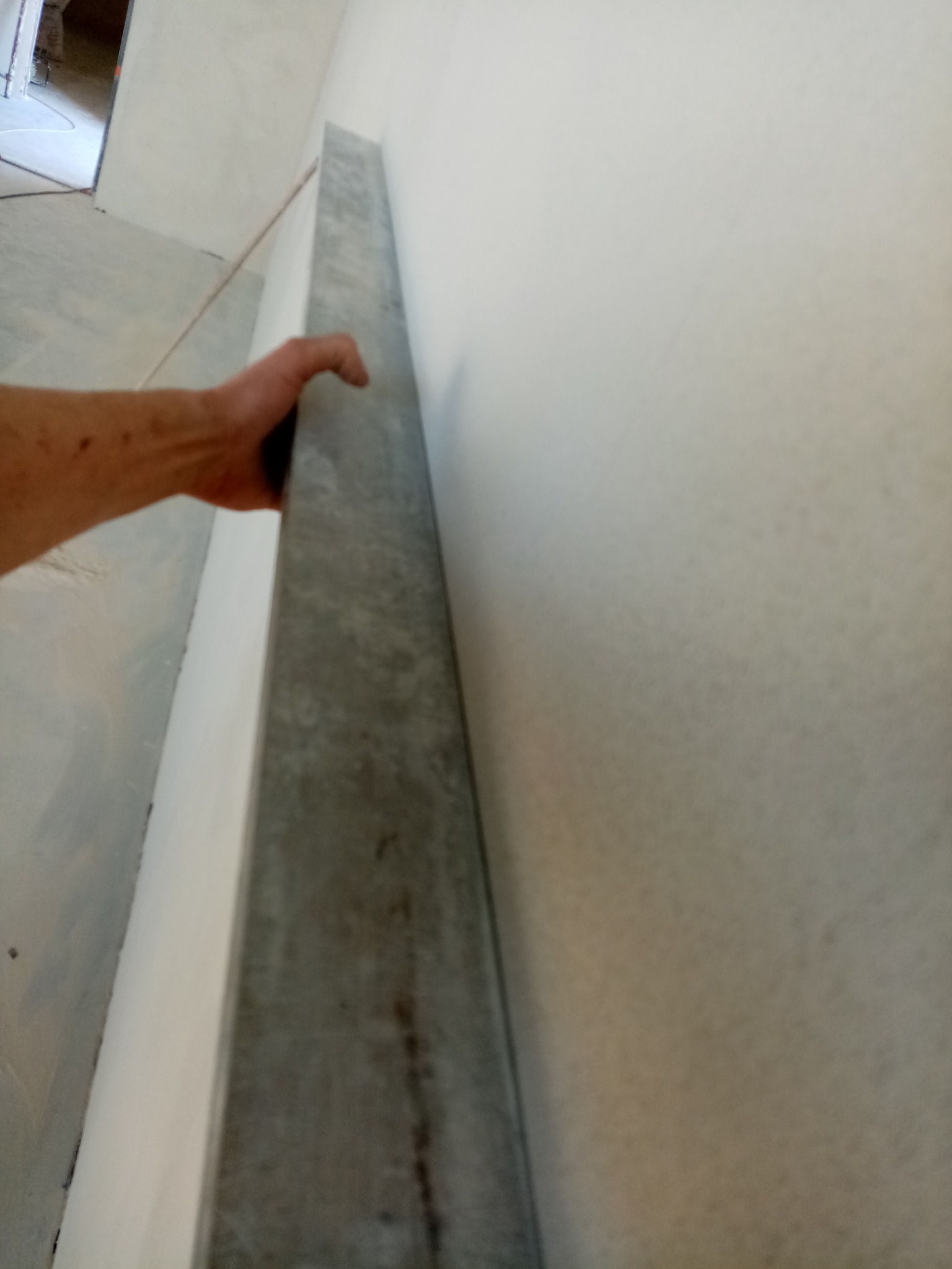 Ręka przytrzymuje metalowy profil przy ścianie przygotowanej do prac wykończeniowych, widoczny fragment pomieszczenia w trakcie remontu.