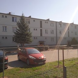 Izol- Complex Tomasz Romanowski - Trzypiętrowy budynek mieszkalny z szarą elewacją, widoczny zza ogrodzenia, z zaparkowanymi samochodami i suszarką na pranie na pierwszym planie.