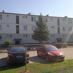 Izol- Complex Tomasz Romanowski - Trzypiętrowy budynek mieszkalny z szarą elewacją, widoczny z poziomu parkingu z dwoma samochodami, przed budynkiem rośnie duża choinka.