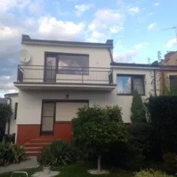 Dwupiętrowy dom z jasną elewacją, balkonem z czarną balustradą i czerwoną ceglaną podstawą, widok z ogrodu z zielonym trawnikiem i drzewami.