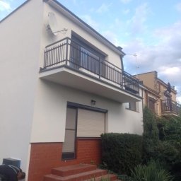 Izol- Complex Tomasz Romanowski - Pracownik maluje dolną część elewacji domu z ceglaną podmurówką, widoczny balkon z czarną balustradą i satelita na ścianie.