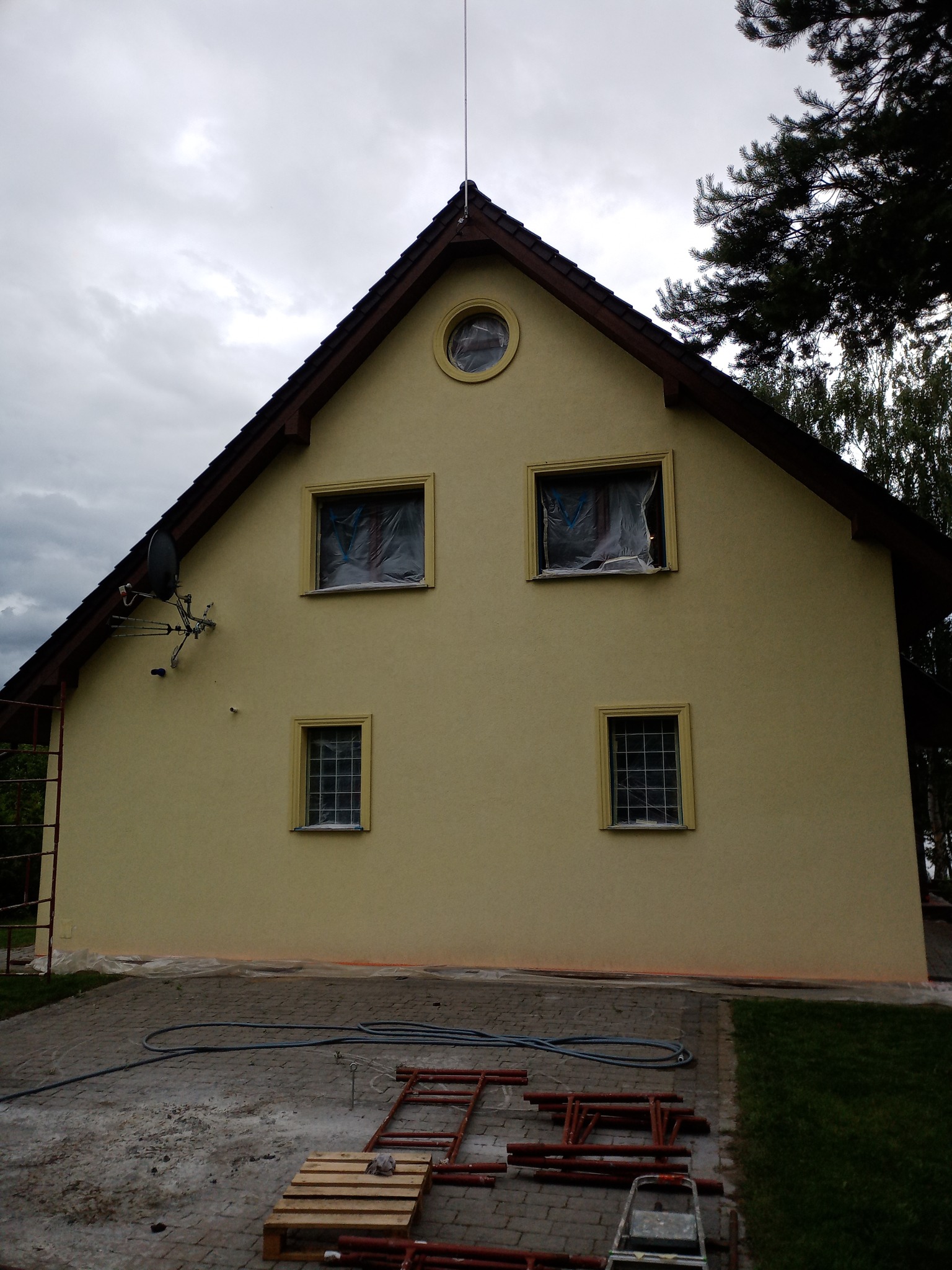 Żółta elewacja domu jednorodzinnego po odświeżeniu, z oknami zabezpieczonymi folią malarską, widoczna paleta i rusztowanie na kostce brukowej.