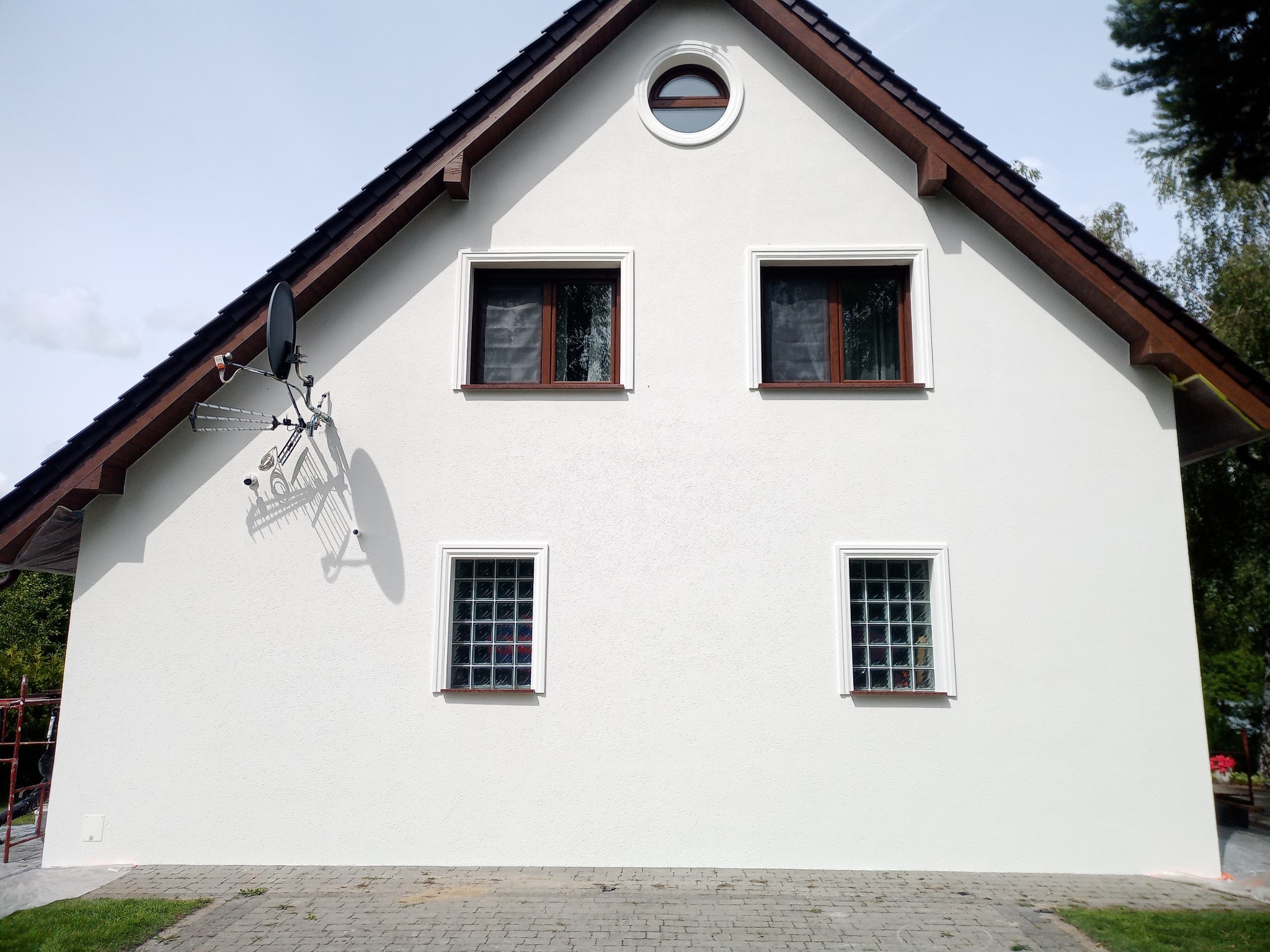 Biała elewacja dwukondygnacyjnego domu z brązowymi obramowaniami okien i dachu, z anteną satelitarną po lewej stronie i okrągłym oknem na szczycie.