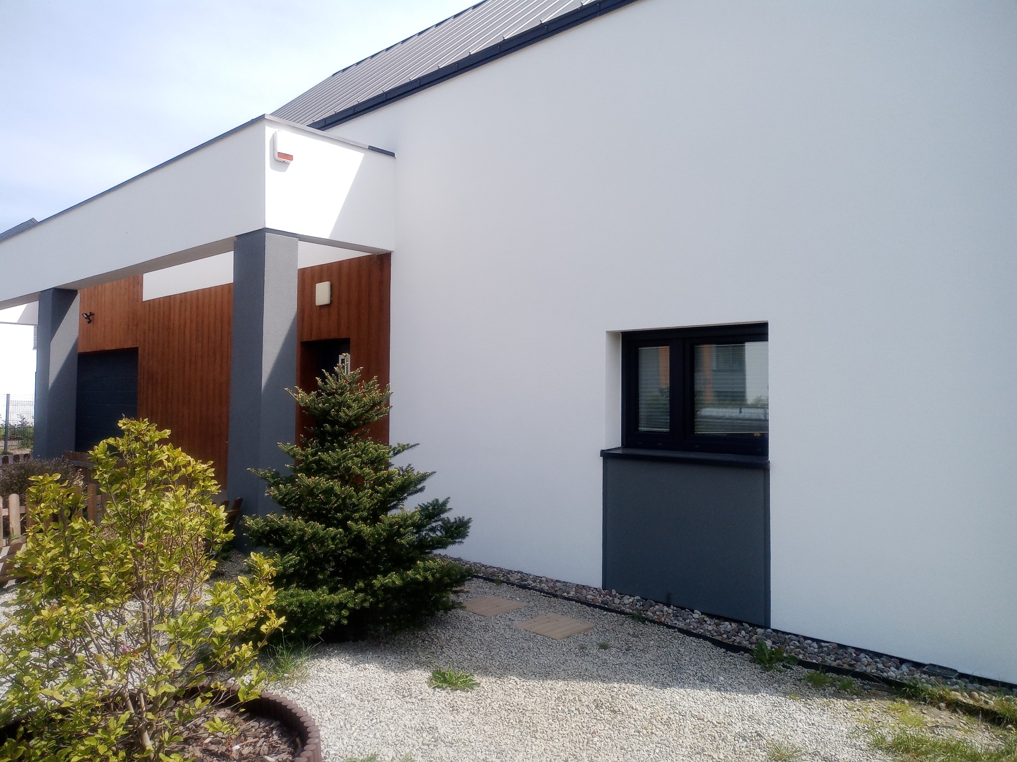 Nowoczesny dom z białą elewacją, szarymi akcentami i minimalistycznym oknem, otoczony ozdobnym żwirem i zielenią.